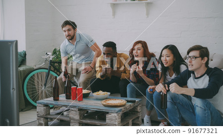 Group of young friends watching olympic games match on TV together eating snacks and drinking beer on holidays at home indoors. Some of them happy with their team winning but others disappointed Group of young friends watching olympic games match on TV together eating snacks and drinking beer on holidays at home indoors. Some of them happy with their team winning but others disappointed 91677321