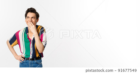 Happy young man laughing and covering mouth with hand, looking up at something funny, standing over white background Happy young man laughing and covering mouth with hand, looking up at something funny, standing over white background 91677549