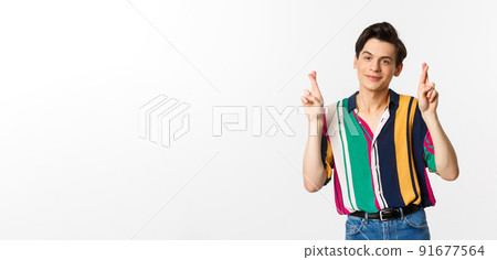 Optimistic young man smiling, holding fingers crossed for good luck, making wish, standing over white background 91677564