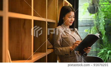 Concentrated young businesswoman standing near bookshelves in bright company office Concentrated young businesswoman standing near bookshelves in bright company office 91678201