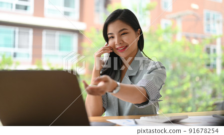 Asian female entrepreneur having conversation on cell phone with her business partner and reading information on laptop Asian female entrepreneur having conversation on cell phone with her business partner and reading information on laptop 91678254