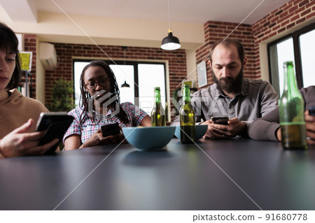 Cultural diverse people sitting in living room while browsing internet on modern smartphone devices. Multiethnic friends sitting at home while watching online video content on mobile phones. Cultural diverse people sitting in living room while browsing internet on modern smartphone devices. Multiethnic friends sitting at home while watching online video content on mobile phones. 91680778