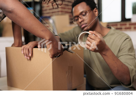 Married partners sealing cardboard boxes with adhesive tape to move furniture in new real estate property. Packing things and shipping storage cargo, enjoying flat relocation. Close up. 91680804