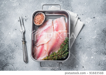 Raw white fish tilapia, uncooked fillet in kitchen tray with herbs. Gray background. Top view 91683588