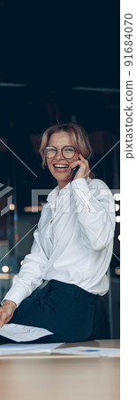 Smiling mature business woman sitting on table and talking on phone in modern office Smiling mature business woman sitting on table and talking on phone in modern office 91684070