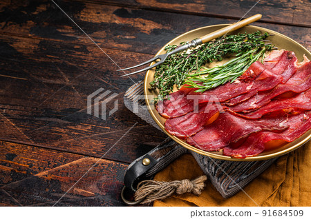 Basturma, sliced dried beef meat, meat Jerky in steel plate with herbs and spices. Wooden background. Top view. Copy space Basturma, sliced dried beef meat, meat Jerky in steel plate with herbs and spices. Wooden background. Top view. Copy space 91684509