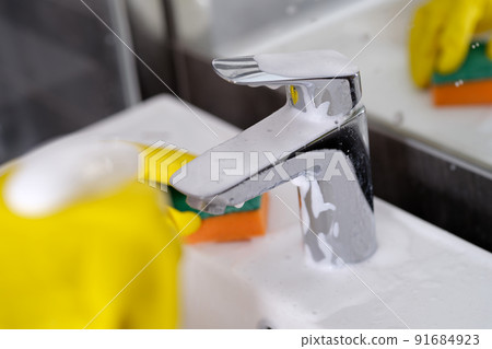 Hand in yellow rubber gloves holding sponge for washing dirty faucet with limescale 91684923