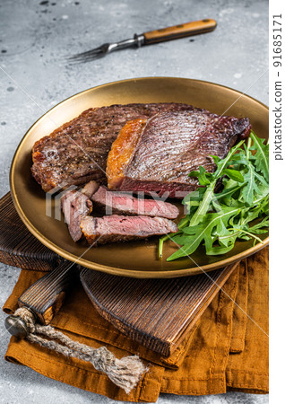 Roast picanha or top sirloin beef meat steak on a plate with arugula salad. Gray background. Top view Roast picanha or top sirloin beef meat steak on a plate with arugula salad. Gray background. Top view 91685171