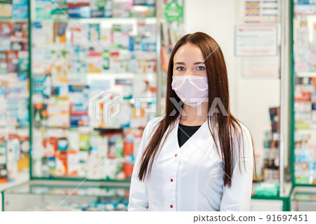 A female pharmacist in a medical mask poses against the background of a pharmacy window in a blur. Concept of virus protection and pharmacy business A female pharmacist in a medical mask poses against the background of a pharmacy window in a blur. Concept of virus protection and pharmacy business 91697451