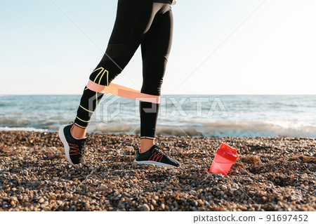 The concept of sport. A young woman exercises with a power band on her legs. There's a pink shaker on the ground. In the background, the sea and the horizon line. Close up The concept of sport. A young woman exercises with a power band on her legs. There's a pink shaker on the ground. In the background, the sea and the horizon line. Close up 91697452