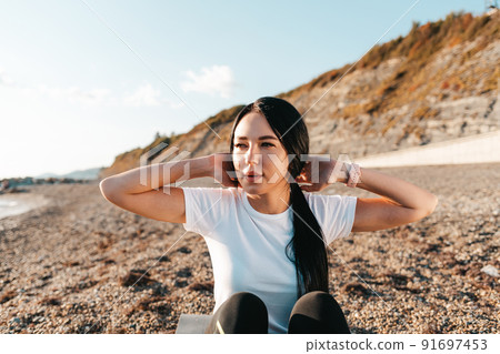 The concept of sport. Portrait of a young brunette woman shakes her abs sitting on a sports Mat. In the background mountains and wild beach The concept of sport. Portrait of a young brunette woman shakes her abs sitting on a sports Mat. In the background mountains and wild beach 91697453