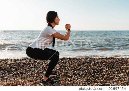The concept of sport. Young brunette woman doing sports and squats. In the background, the sea and the shore. Copy space The concept of sport. Young brunette woman doing sports and squats. In the background, the sea and the shore. Copy space 91697454