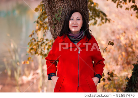 Portrait of beautiful adult smiling woman in a red coat posing by a tree. Autumn season 91697476