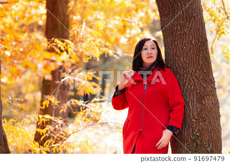 Elegant beautiful mature woman in a red coat posing by a tree. Autumn season. Copy space Elegant beautiful mature woman in a red coat posing by a tree. Autumn season. Copy space 91697479