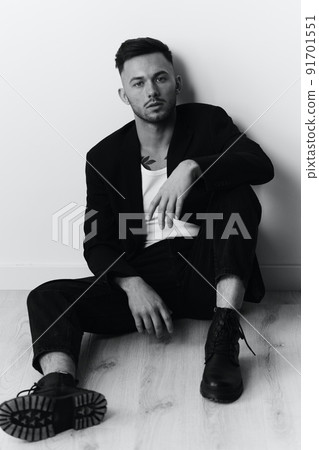 Modelling snapshots. Serious self-confident attractive handsome man in black jacket sitting on floor posing in white studio background. Black and White concept. Copy space 91701551