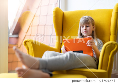 Portrait of cute adorable blond little kid girl enjoy sitting in cozy soft yellow armchair playing game or watching TV show movies using tablet. Children smart technology gadget usage concept Portrait of cute adorable blond little kid girl enjoy sitting in cozy soft yellow armchair playing game or watching TV show movies using tablet. Children smart technology gadget usage concept 91705271