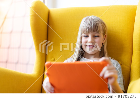 Portrait of cute adorable blond little kid girl enjoy sitting in cozy soft yellow armchair playing game or watching TV show movies using tablet. Children smart technology gadget usage concept Portrait of cute adorable blond little kid girl enjoy sitting in cozy soft yellow armchair playing game or watching TV show movies using tablet. Children smart technology gadget usage concept 91705273