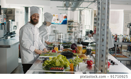 Portrait of man in uniform preparing green salad on cutting board to cook gastronomy cuisine dish for restaurant menu. Professional chef working on culinary recipe with organic food. Handheld shot. 91710925
