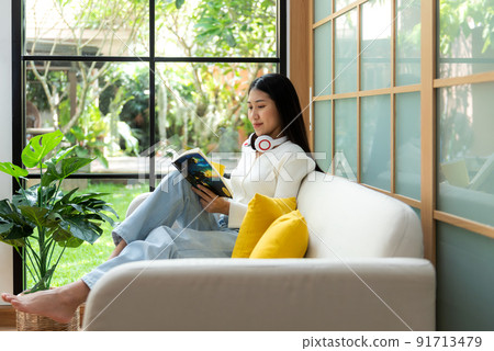 Happy young woman reading book on sofa at home.  Lifestyle freelance relax and chill drinking coffee  91713479