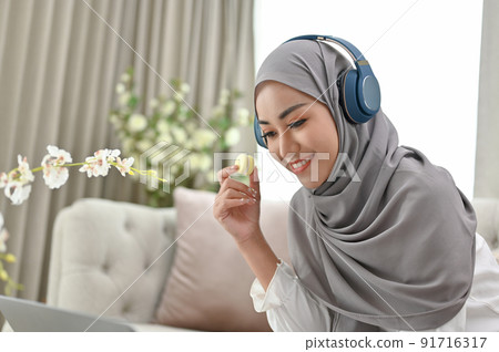 Attractive millennial Asian Muslim woman eating macaron while using laptop 91716317
