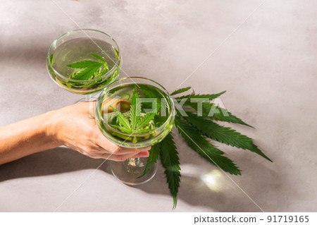 A woman's hand holds glass with green drink from SBD hemp, light concrete background hard shadows A woman's hand holds glass with green drink from SBD hemp, light concrete background hard shadows 91719165