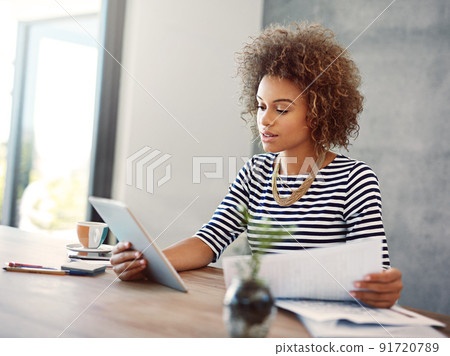 Comparing figures. Shot of a young woman a digital tablet and reading paperwork while working at home. Comparing figures. Shot of a young woman a digital tablet and reading paperwork while working at home. 91720789