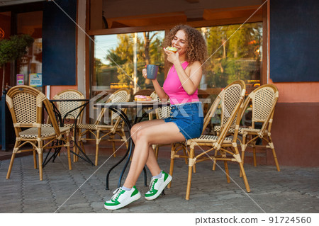 young attractive curly woman sits at table in cafe on summer terrace. Colorful delicious donuts, sweet pastries. Good mood, smiling. Rest, snack break. Enjoys young attractive curly woman sits at table in cafe on summer terrace. Colorful delicious donuts, sweet pastries. Good mood, smiling. Rest, snack break. Enjoys 91724560