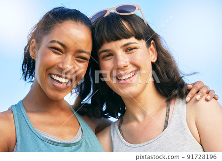 Portrait of two smiling friends embracing while standing together against blue sky. Smiling happy young women bonding and hugging outside over summer break. Close best friends having fun on a weekend 91728196