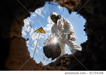 View from inside pit of ecologist digging pit by shovel and planting tree, wearing gas mask, protective overalls outdoors, standing near biohazard symbol warning about dangerous biological materials. 91734298