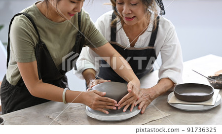Young woman teaching mature woman making ceramic pot from clay in pottery workshop. Activity, handicraft, hobbies concept Young woman teaching mature woman making ceramic pot from clay in pottery workshop. Activity, handicraft, hobbies concept 91736325