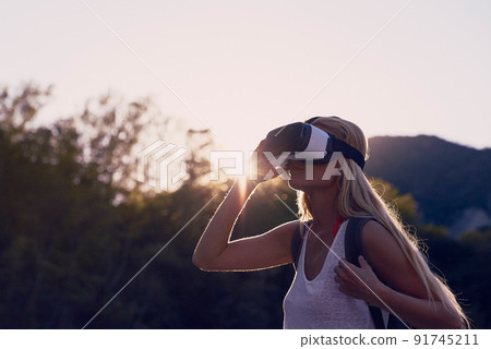 Woman hiking at sunrise using VR goggles to explore the metaverse. Blond woman on holiday hiking in a forest using VR goggles to experience augmented virtual reality 91745211