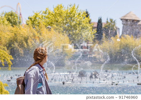 A woman with pigtails looks at a fountain spouting from a lake against the backdrop of sun-drenched trees and park architecture. Weekend at the amusement park. 91746960