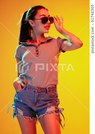 Portrait of stylish young girl posing in casual outfit and sunglasses isolated over yellow background in neon light 91748305