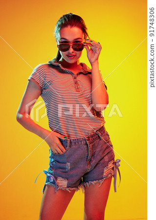 Portrait of beautiful young girl posing in casual outfit and sunglasses isolated over yellow background in neon light Portrait of beautiful young girl posing in casual outfit and sunglasses isolated over yellow background in neon light 91748306