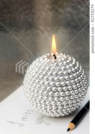 Close-up of beautiful round silver candle burning on sheet of paper with inscription I miss you, hand lettering. Against the background of frosted window glass at night. Letter memories. Black pencil 91750274