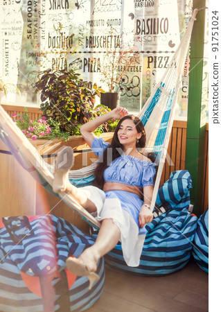 A beautiful young woman lies in a hammock on the summer terrace of a city cafe and enjoys the summer A beautiful young woman lies in a hammock on the summer terrace of a city cafe and enjoys the summer 91751024
