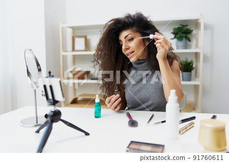 Makeup artist. Lovely tanned handsome curly Latin beauty blogger in casual top present serum for skin rejuvenation in home interior. Copy space. Influencer record video review blog using smartphone Makeup artist. Lovely tanned handsome curly Latin beauty blogger in casual top present serum for skin rejuvenation in home interior. Copy space. Influencer record video review blog using smartphone 91760611