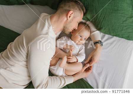 A loving father lies on the bed with his baby son and hugs him A loving father lies on the bed with his baby son and hugs him 91771846