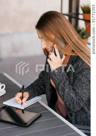 A young charming woman is calling on a mobile phone, sitting in a cafe and talking on a mobile phone with a smile, writing notes in a notebook. Remote work 91772303