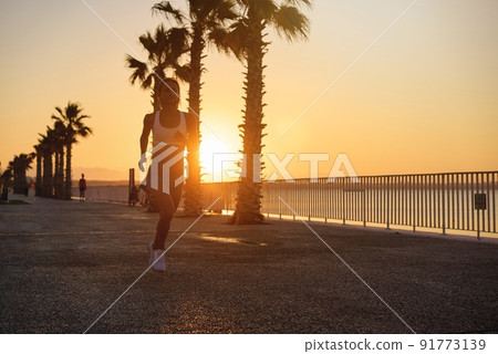 Silhouette of young woman jogging on shore at sunrise. 91773139
