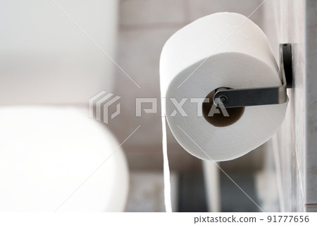 Roll of toilet paper hanging on a metallic holder on a bathroom wall. 91777656