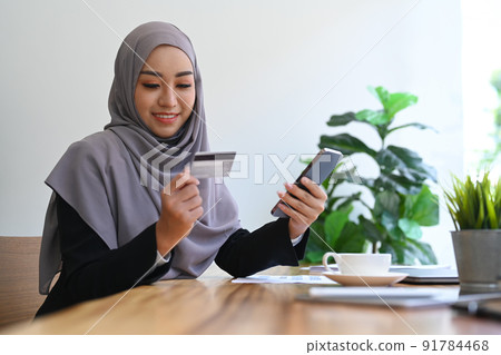 Smiling muslim woman holding credit card and using smart phone, purchasing product via online shopping 91784468