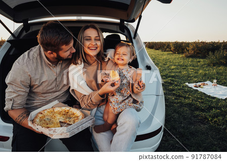 Happy Young Family Sitting in the Trunk of SUV Car and Eating Pizza, Mom Dad and Baby Daughter Having Fun Time on Weekend Outdoors Happy Young Family Sitting in the Trunk of SUV Car and Eating Pizza, Mom Dad and Baby Daughter Having Fun Time on Weekend Outdoors 91787084