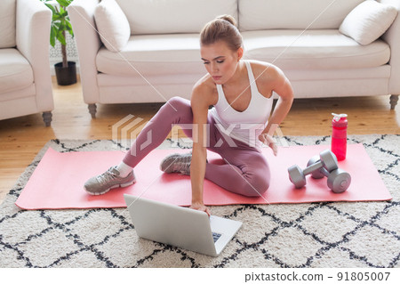 Cheerful slim sporty woman sitting on the mat with dumbbells and bottle of water 91805007