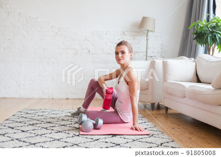 Beautiful slim woman relaxing and drinking proteine shake or water after fitness exercises at home 91805008