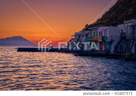 Sunset in fishermen village of Klima, Milos island, Greece. Pair of adults enjoy sunset view on pier of small fishermen's village. Sunset in fishermen village of Klima, Milos island, Greece. Pair of adults enjoy sunset view on pier of small fishermen's village. 91807371