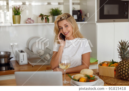 Blonde cheerful young woman talking on cellphone at laptop in kitchen with white wine. 91815418