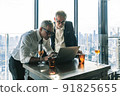 Picture of two business men negotiating over a deal. One man is in his 70th he is wearing black suit. The other man is in his 40th he is wearing a white shirt and black tie. They sat in lounge. 91825655
