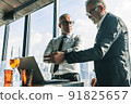 Picture of two business men negotiating over a deal. One man is in his 70th he is wearing black suit. The other man is in his 40th he is wearing a white shirt and black tie. They sat in lounge. 91825657