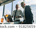 Picture of two business men negotiating over a deal. One man is in his 70th he is wearing black suit. The other man is in his 40th he is wearing a white shirt and black tie. They sat in lounge. 91825659